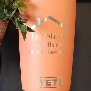 May include: A coral-colored Yeti tumbler with a silver lid and base. The tumbler features the text "That's What I Do, I Sell Homes, I Drink & I Know Things" and a house graphic. The Yeti logo is visible.
