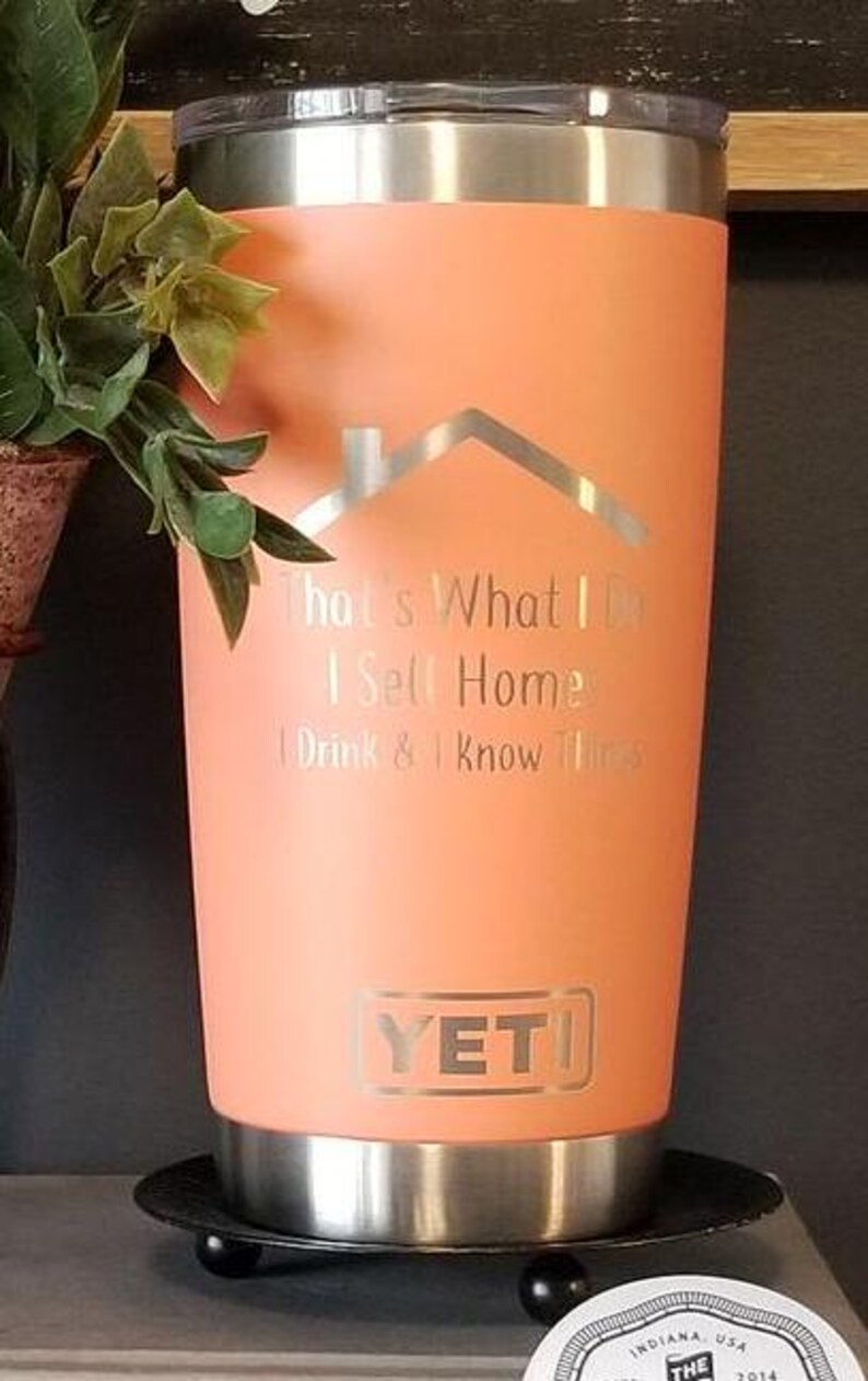 May include: A coral-colored Yeti tumbler with a silver lid and base. The tumbler features the text "That's What I Do, I Sell Homes, I Drink & I Know Things" and a house graphic. The Yeti logo is visible.