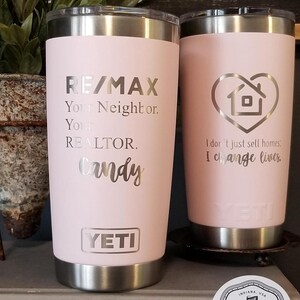 May include: Two pink insulated tumblers with silver accents. One features "RE/MAX" and "Candy" text, the other has a house-shaped heart and the text "I don't just sell homes, I change lives." Both have the "YETI" logo.