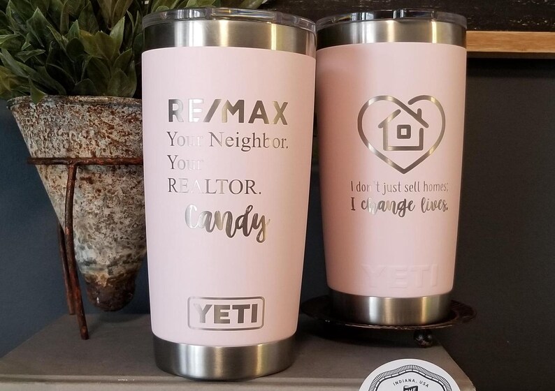 May include: Two pink insulated tumblers with silver accents. One features "RE/MAX" and "Candy" text, the other has a house-shaped heart and the text "I don't just sell homes, I change lives." Both have the "YETI" logo.