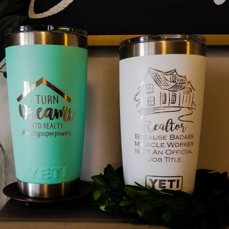 May include: Two Yeti tumblers with silver rims. One is teal with the text "Turn Dreams to Reality" and a house graphic. The other is white with a house graphic and the text "Realtor Because Badass Miracle Worker Isn't an Official Job Title."
