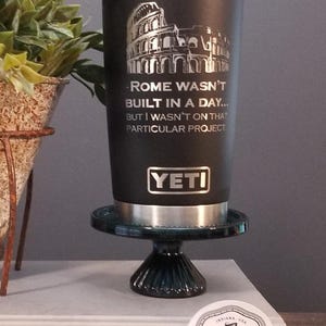 May include: Black Yeti tumbler with a silver rim and lid, featuring an engraved image of the Colosseum and the text "Rome Wasn't Built in a Day... But I Wasn't on That Particular Project." The tumbler is displayed on a small, dark green pedestal.