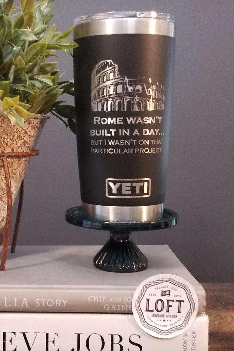 May include: Black Yeti tumbler with a silver rim and lid, featuring an engraved image of the Colosseum and the text "Rome Wasn't Built in a Day... But I Wasn't on That Particular Project." The tumbler is displayed on a small, dark green pedestal.