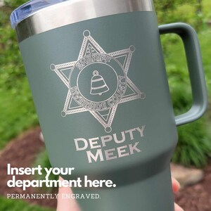 May include: A green stainless steel travel mug with a silver engraved sheriff's star badge. The badge has the words "Sheriff Riverside County" and a bell inside. The text "Deputy Meek" is engraved below the badge.