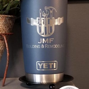 May include: A navy blue YETI tumbler with a silver engraved design of tools and the initials "JMF". The text "JMF BUILDING & REMODELING" is below the design. The tumbler sits on a black coaster.