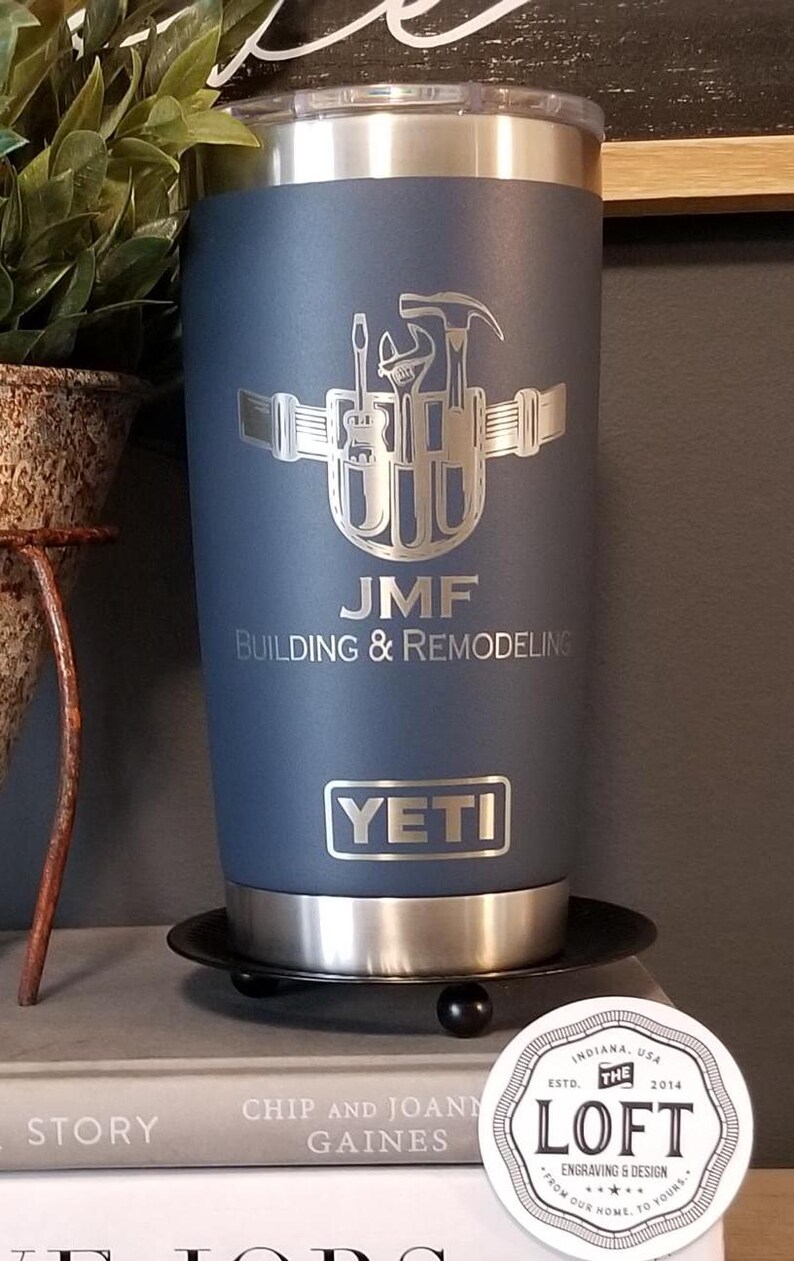 May include: A navy blue YETI tumbler with a silver engraved design of tools and the initials "JMF". The text "JMF BUILDING & REMODELING" is below the design. The tumbler sits on a black coaster.