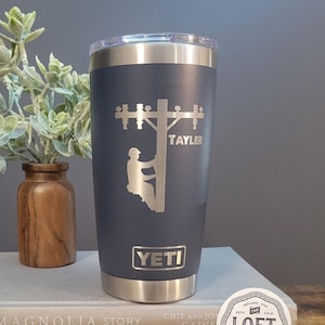 May include: A dark blue YETI tumbler with a stainless steel lid and bottom. The tumbler has a laser engraved design of a lineman climbing a utility pole with the name "TAYLER" engraved below.