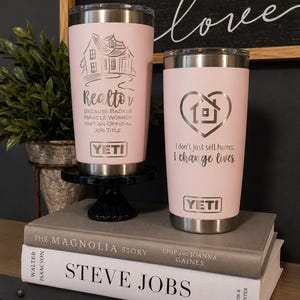 May include: Two light pink stainless steel YETI tumblers with silver laser-engraved text and graphics. The left tumbler features a house illustration and the text Realtor BECAUSE BADASS MIRACLE WORKER ISN'T AN OFFICIAL JOB TITLE. The right tumbler shows a heart-shaped house icon and the text I don't just sell homes; I change lives.