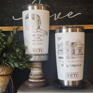 May include: Two white YETI tumblers with silver accents. One features a tape measure graphic and the text "KEEP CALM AND BUILD ON JED SISK." The other has a house illustration and the text "Turn your dreams into reality, Cay Ponzurich."