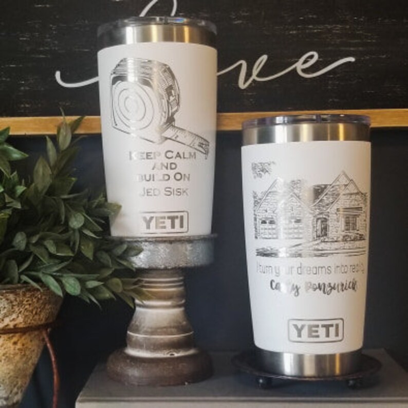 May include: Two white YETI tumblers with silver accents. One features a tape measure graphic and the text "KEEP CALM AND BUILD ON JED SISK." The other has a house illustration and the text "Turn your dreams into reality, Cay Ponzurich."