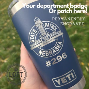 May include: A blue Yeti tumbler with a silver lid and a silver engraved design. The design features a building, the words "State Patrol Nebraska", and the number "296". The text "Your department badge or patch here! Permanently engraved." is at the top of the tumbler.