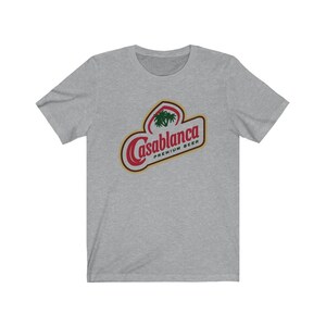 May include: A heather grey t-shirt with a red and gold logo that says "Casablanca Premium Beer". The logo features a palm tree.