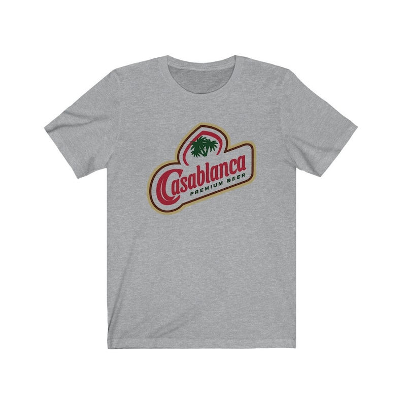 Op de afbeelding: Een heidekleurig t-shirt met een rood en gouden logo met de tekst "Casablanca Premium Beer". Het logo bevat een palmboom.