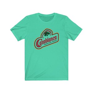 Op de afbeelding: Een turquoise blauw t-shirt met een rood en gouden logo met de tekst "Casablanca Premium Beer" met een palmboom afbeelding.