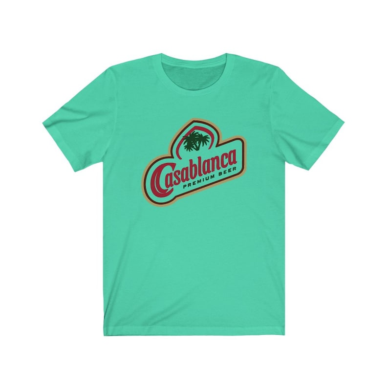 Op de afbeelding: Een turquoise blauw t-shirt met een rood en gouden logo met de tekst "Casablanca Premium Beer" met een palmboom afbeelding.