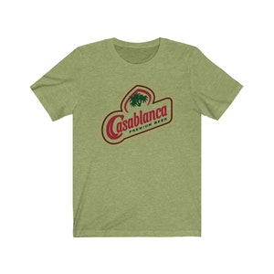 Op de afbeelding: Een groen T-shirt met een rood en wit logo met de tekst "Casablanca Premium Beer". Het logo bevat een palmboom.