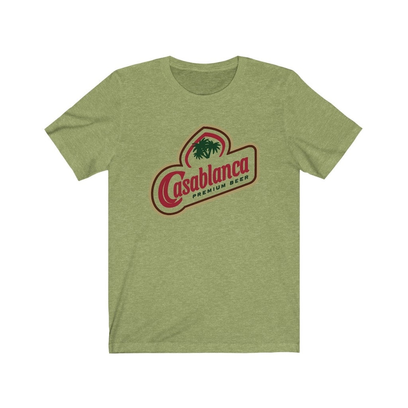 Op de afbeelding: Een groen T-shirt met een rood en wit logo met de tekst "Casablanca Premium Beer". Het logo bevat een palmboom.