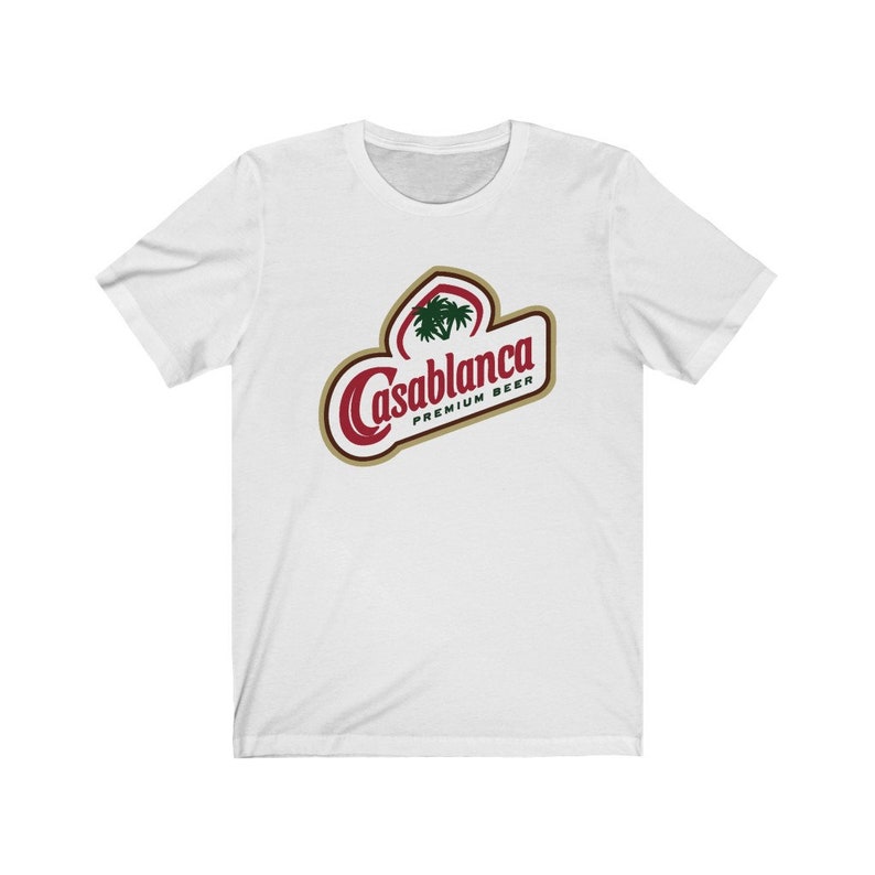 Op de afbeelding: Wit T-shirt met een rood en gouden logo met de tekst "Casablanca Premium Beer". Het logo bevat een gestileerde palmboom.