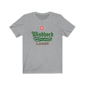 May include: A heather grey t-shirt with a green and gold Windhoek Lager logo. The logo features a stylized 'W' in a red shield and the text 'Windhoek Lager' in a green script font.