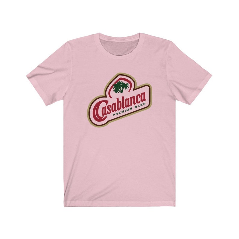 Op de afbeelding: Roze T-shirt met een rood en gouden logo met de tekst "Casablanca Premium Beer". Het logo bevat een palmboom.