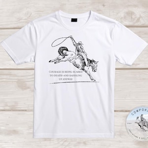 May include: White t-shirt featuring a black and white illustration of a cowboy riding a bucking horse. The text "COURAGE IS BEING SCARED TO DEATH-AND SADDLING UP ANYWAY" is printed below the image. A circular logo is in the bottom right corner.