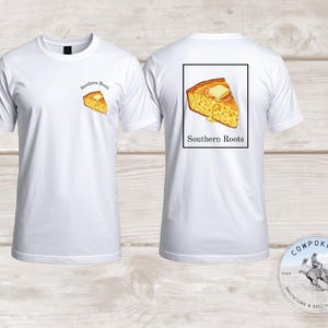 Puede incluir: Camiseta blanca con un diseño de rebanada de pan de maíz con mantequilla y el texto "Southern Roots". El mismo diseño está en la parte posterior, dentro de un rectángulo negro. Pequeño logotipo en la parte inferior derecha.