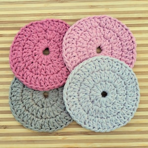 May include: Four crocheted coasters in pink and gray. The coasters are round and have a hole in the center. They are arranged in a stack on a wooden surface.