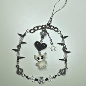 May include: A phone charm featuring a silver chain adorned with metal spikes, clear beads, and small silver spheres. A central pendant displays a black heart above a translucent white alien figure, accompanied by a small silver star charm.