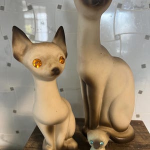 May include: Three ceramic cat figurines. The largest is a Siamese cat with blue eyes. The other two are smaller, one with orange eyes and the other with blue eyes. All are in a sitting position and have a vintage aesthetic.