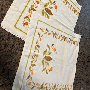 May include: Two rectangular off-white fabric napkins featuring a printed border of orange fruit and brown and green leaves. The design includes a thin olive green line framing the botanical pattern, which is repeated on both pieces of textile.