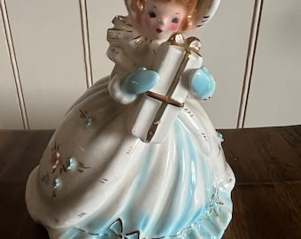 May include: A porcelain figurine of a girl wearing a white dress with blue trim and a white hat. She is holding a white gift box with a gold ribbon. The figurine has a vintage look and is decorated with floral details.