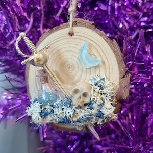 May include: A rustic wood slice ornament with a miniature skull, sword, and potion bottle. The ornament features a crescent moon, blue and white moss, and a natural twine hanger. The background is a purple tinsel.