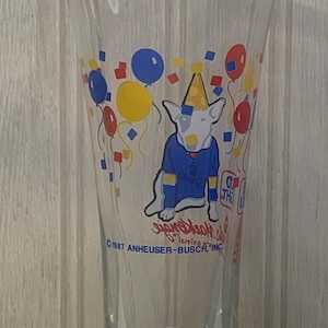 May include: A clear glass beer mug with a colorful cartoon dog wearing a party hat and surrounded by balloons. The mug has the text "Budweiser" and "1987 Anheuser-Busch, Inc." printed on it.