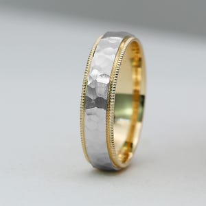 May include: A two-tone wedding band with a hammered silver center and a gold-colored edge. The ring has a textured surface and a beaded border. The band is a classic design for a wedding or engagement.