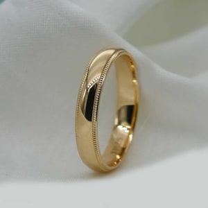 May include: A gold wedding band with a polished finish. The ring features a detailed, textured edge. The band is set against a soft, white background, highlighting its reflective surface and classic design.