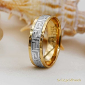 May include: A gold and white wedding band with a Greek key design. The band is about 6mm wide and has a polished finish.
