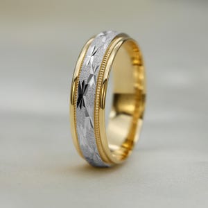 May include: A gold wedding band with a silver geometric pattern in the centre. The ring has a polished finish and a textured design. The band is approximately 6mm wide.