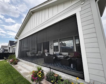 Peut inclure: Vue extérieure d'une maison avec une véranda grillagée. La véranda est équipée de moustiquaires noires et de meubles de jardin. La maison a un bardage blanc et une pelouse verte avec des parterres de fleurs au premier plan.