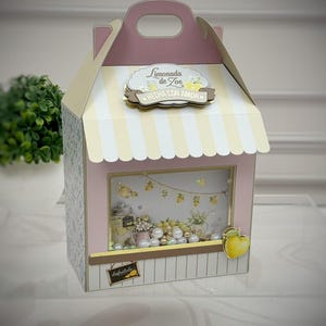 May include: A pink and cream paper gift box shaped like a small shop with a striped awning. The front features a clear window displaying lemon-themed decorations, white pearls, and a sign reading Limonada de Zoe HECHA CON AMOR and disfrútalo.