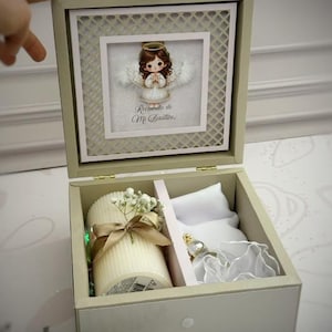 Può includere: Una scatola ricordo beige con coperchio incernierato decorata con l'illustrazione di un angelo e la scritta "Recuerdo de Mi Bautizo". All'interno, una candela color crema a coste con nastro dorato e gipsofila accanto a un sacchetto in raso bianco.