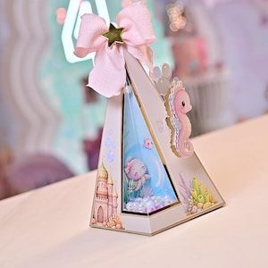 May include: A decorative, triangular box with a mermaid theme. The box features a pink bow with a gold star, a seahorse, and illustrations of a mermaid, castle, and underwater scene. The box is white with gold trim.