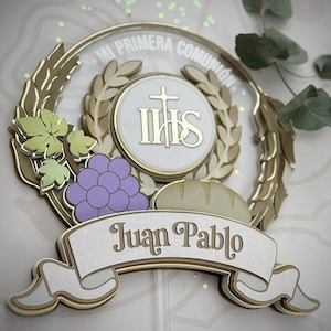 Pode incluir: Topper de bolo para a Primeira Comunhão. O design inclui uma coroa dourada, uma faixa com o nome "Juan Pablo", uma cruz, uvas e pão. O texto "MI PRIMERA COMUNIÓN" é visível.