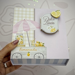 May include: A rectangular box with a light purple and white striped base. The front features a graphic of a lemonade cart with a pink and white umbrella, a jar of lemonade, and lemons. A label on the box reads Dulce Limón with a heart-shaped lemon.