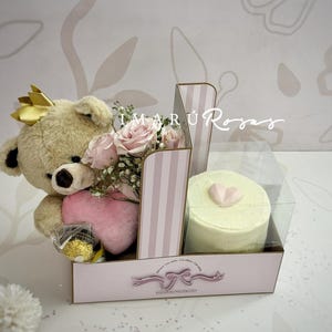 Puede incluir: Caja de regalo con un oso de peluche con corona dorada, rosas rosas, una almohada en forma de corazón y un pastel pequeño con decoración en forma de corazón. La caja tiene divisores a rayas rosas y blancas.