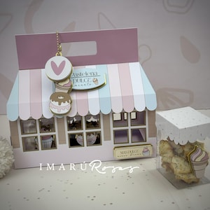 Könnte beinhalten: Eine dekorative Geschenkbox im Stil einer Konditorei mit Griff, mit einem rosa-blau gestreiften Vordach und Fenstern. Daneben eine transparente Box mit Keksen und einer dekorativen Eistüte. Auf der Box steht der Text "Pastelena Dulce Encanto".
