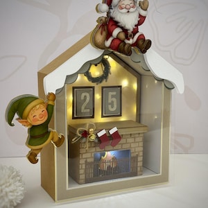 Puede incluir: Una caja de sombras con temática navideña con una chimenea en miniatura, calcetines y la fecha "25" mostrada. Papá Noel y un elfo están posados en el techo y en el lateral. La caja es de cartón y tiene un techo blanco.