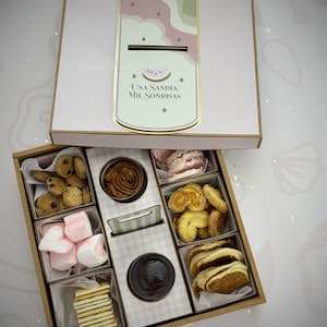 Pode incluir: Uma caixa de presente quadrada com uma tampa apresentando um gráfico de melancia e o texto "UNA SANDIA, MIL SONRISAS". A caixa contém seis compartimentos preenchidos com lanches variados, incluindo biscoitos, marshmallows, bolachas, doces e dois pequenos potes de cremes.