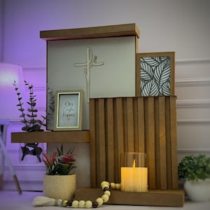 Puede incluir: Un expositor de madera de varios niveles con un panel central con una cruz recortada, un marco pequeño con el texto Ora Confia Espera, un panel con patrón de hojas, un portavelas de cristal con vela encendida y cuentas de madera.