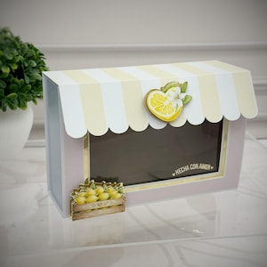 May include: A rectangular box designed like a small shop with a light yellow and white striped awning. A heart-shaped lemon slice with a white flower is attached to the awning. A small crate of lemons is on the front, and the text HECHA CON AMOR is printed.