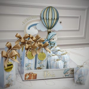 Puede incluir: Exhibición de recuerdos de baby shower con temática de osito de peluche. Los recuerdos están en cajas y bolsas de cuadros azules y blancos, adornados con lazos y etiquetas doradas. También hay un globo aerostático y una figura de osito de peluche.